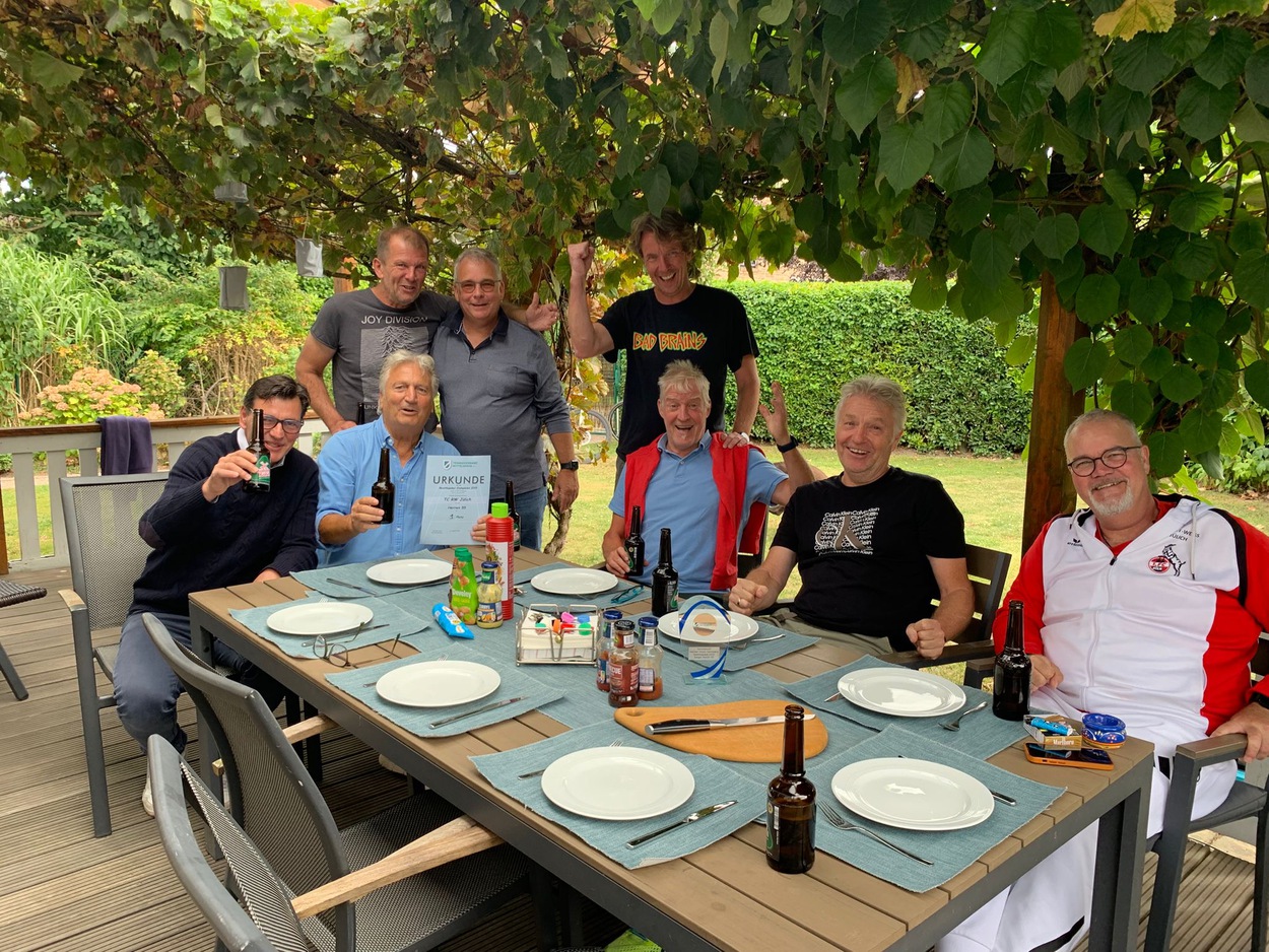 Acht Männer sitzen und stehen an einem gedeckten Tisch in einem Garten